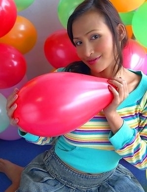 Teaza Tsing stands in front of balloons and she begins playing with them. She has on a colourful top and a jean skirt.